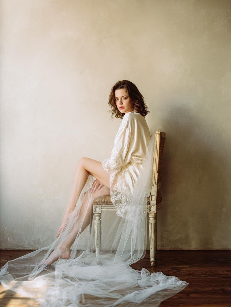 Bride in silk robe seated on a vintage chair with cathedral veil, editorial wedding portrait