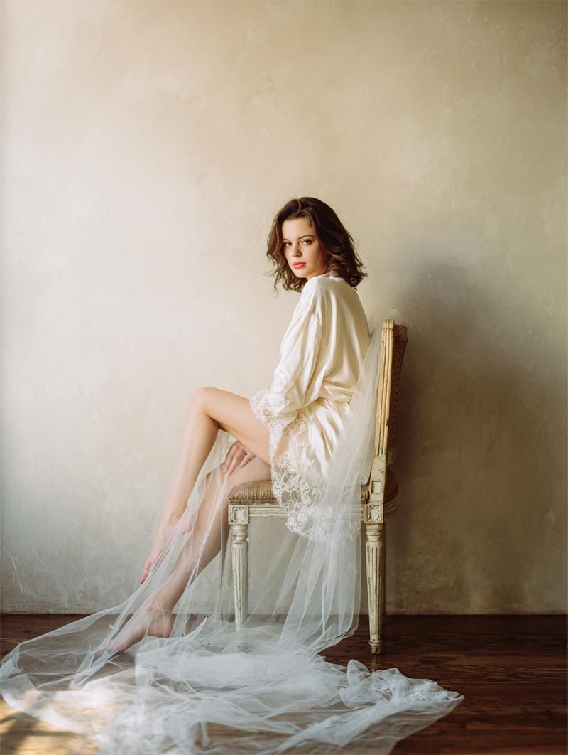 Bride in a flowing ivory silk robe seated on an antique French chair with her cathedral veil draping across hardwood floors