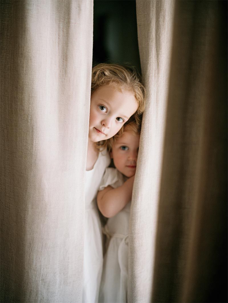Two flower girls in white linen dresses peeking through heavy drapes before a luxury wedding ceremony